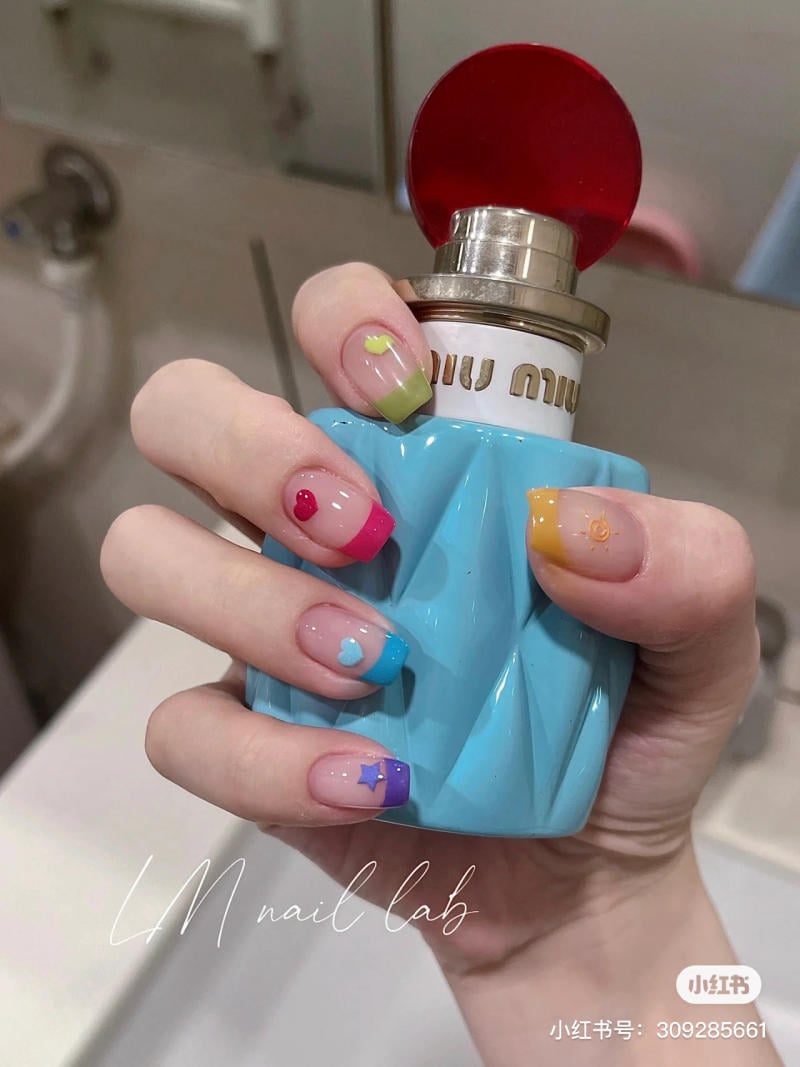Bàn tay với móng tay sơn nhiều màu sắc cầm chai nước hoa màu xanh, nắp đỏ.