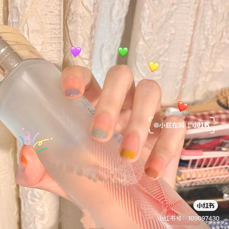 Bàn tay với móng tay sơn nhiều màu khác nhau cầm chai nước, nền có các biểu tượng trái tim nhiều màu.
