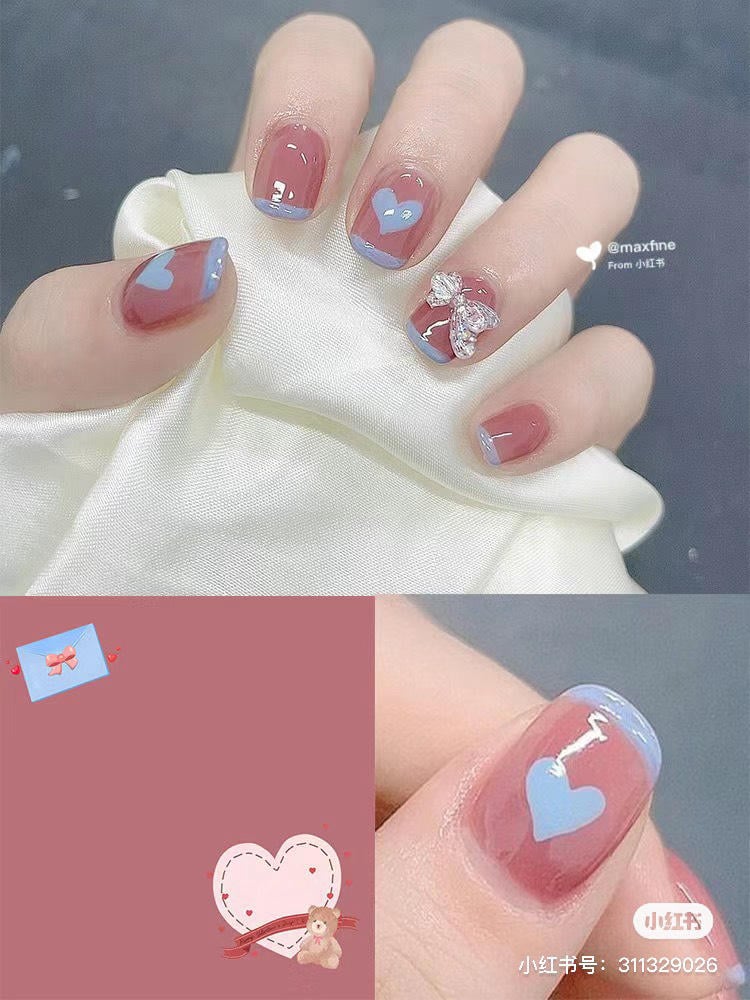 Bàn tay với móng tay sơn màu hồng và xanh, trang trí hình trái tim và nơ bạc, trên nền vải trắng.