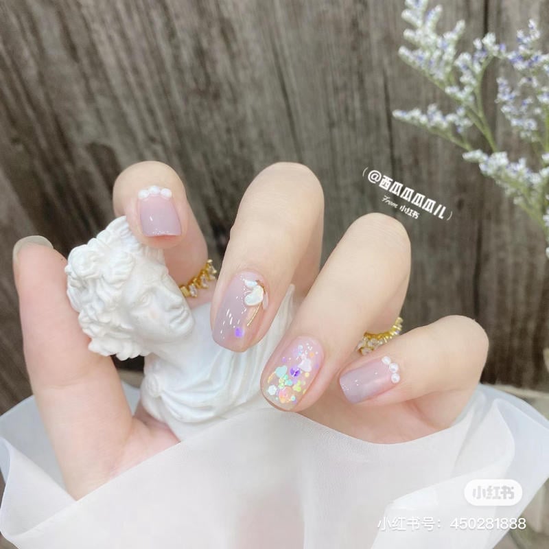Bàn tay với móng tay được trang trí tinh tế, cầm một bức tượng nhỏ, nền gỗ và hoa trang trí bên cạnh.