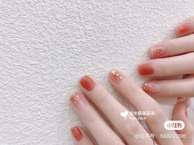 Bàn tay với móng tay màu nâu cam, trang trí lấp lánh vàng, đặt trên nền tường trắng.