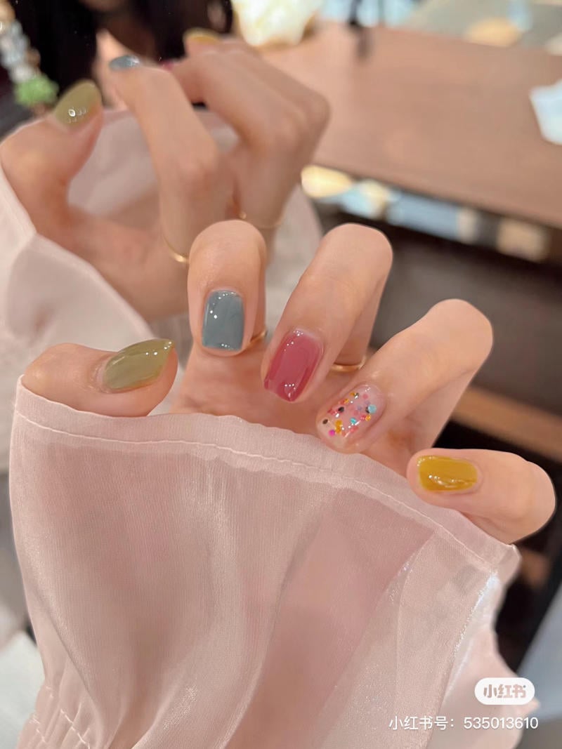 Bàn tay với móng sơn đủ màu sắc, bao gồm xanh, hồng, vàng và một ngón có hạt kim tuyến nhiều màu.