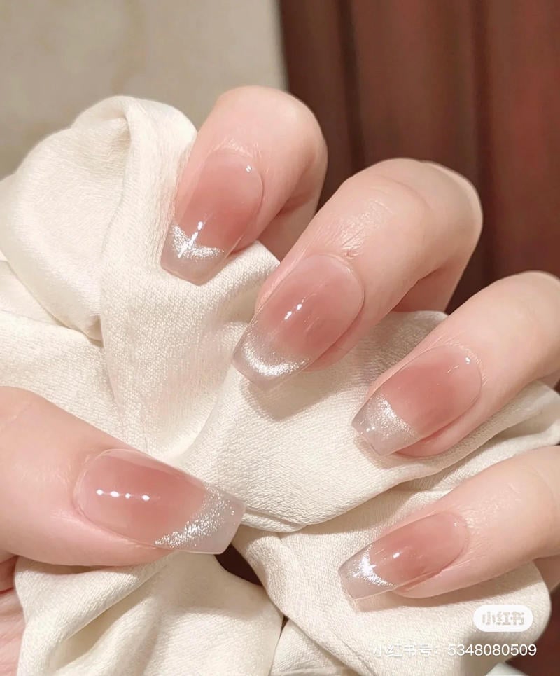 Bàn tay với móng tay sơn màu hồng nhạt, viền trắng lấp lánh, cầm một miếng vải màu trắng.