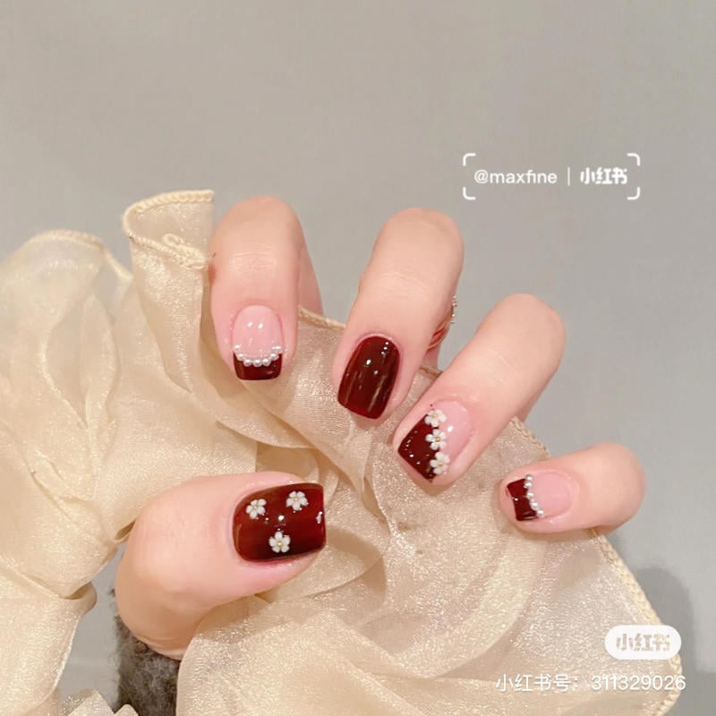 Bàn tay với móng tay sơn màu đỏ và hồng, trang trí hoa và đá, cầm trên nền vải màu vàng nhạt.