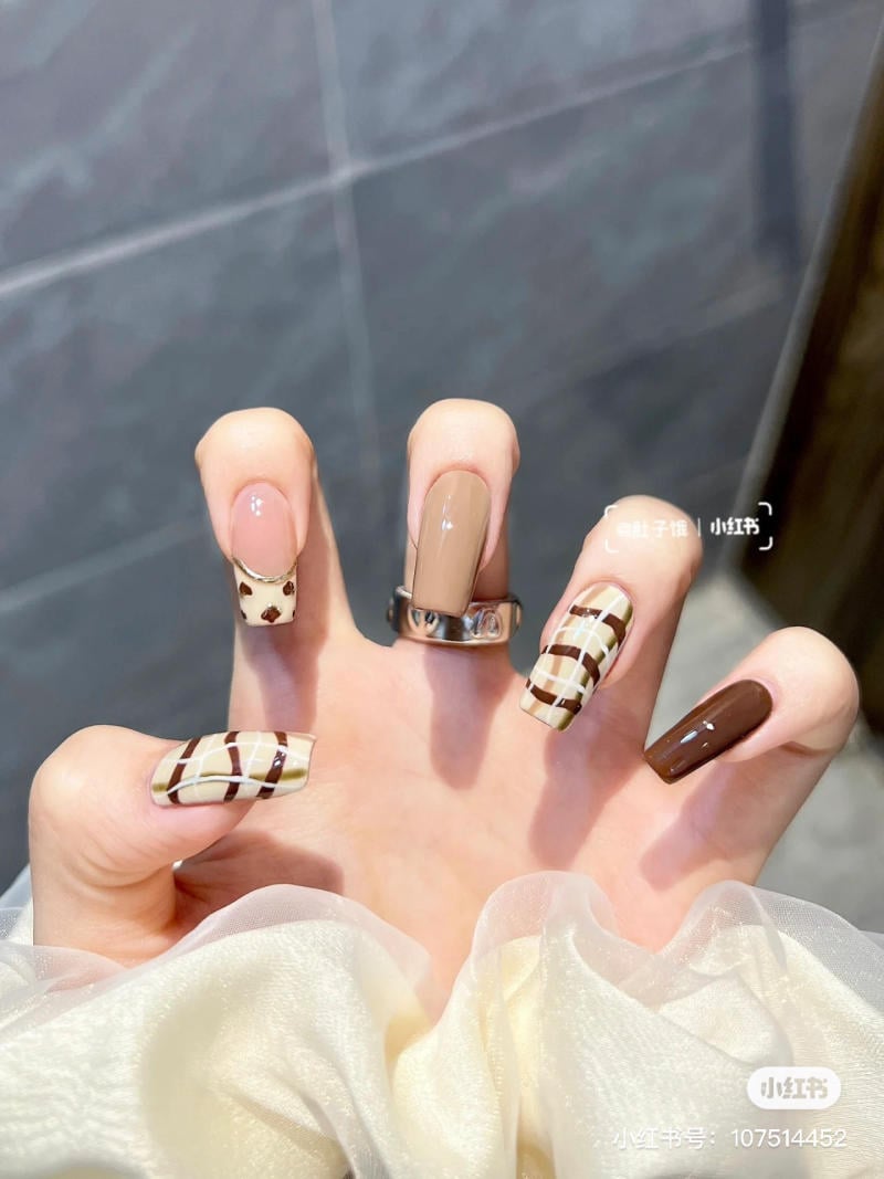 Bàn tay với móng tay được sơn nghệ thuật, họa tiết đa dạng màu nâu và trắng, đeo nhẫn bạc.