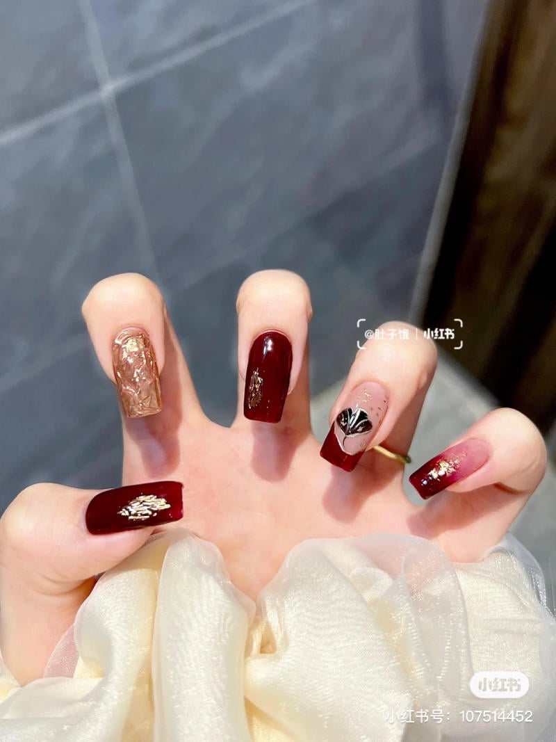 Bàn tay với móng tay sơn màu đỏ, trang trí hoa văn và họa tiết tinh tế, trên nền vải màu sáng.