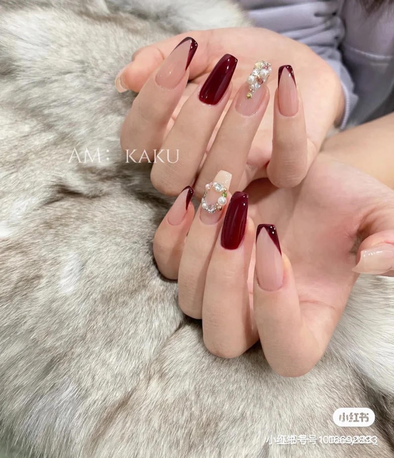 Bàn tay với móng tay dài sơn màu đỏ và nude, một số móng được trang trí với đá và ngọc trai.
