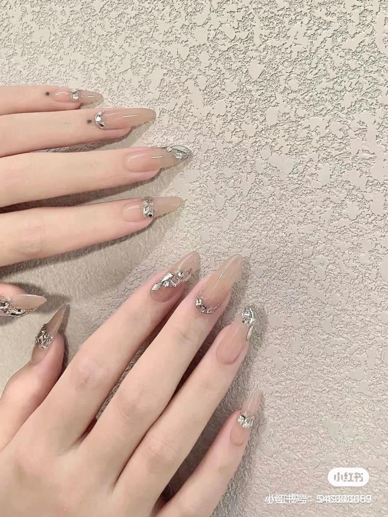 Bàn tay với móng tay dài được trang trí họa tiết bạc, đặt trên nền tường có kết cấu thô.