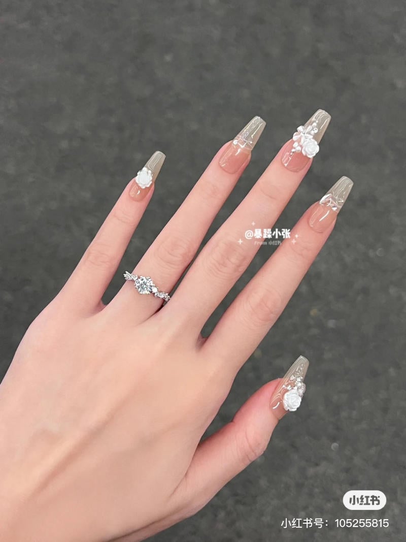 Bàn tay với móng tay dài, trang trí hoa và đá, đeo nhẫn kim cương trên nền xám.