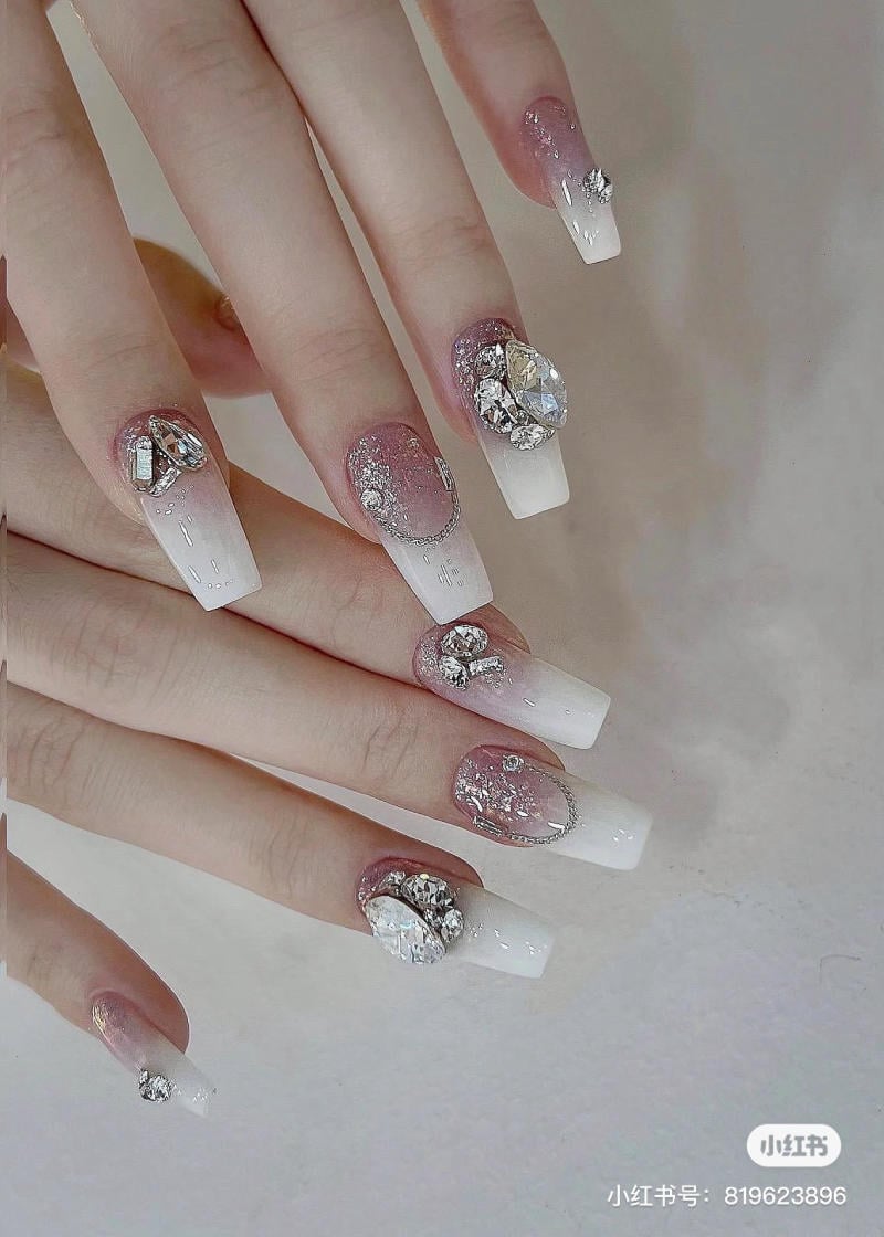 Móng tay trang trí với đá lấp lánh, màu trắng và hồng nhạt, kiểu dáng dài và cầu kỳ.