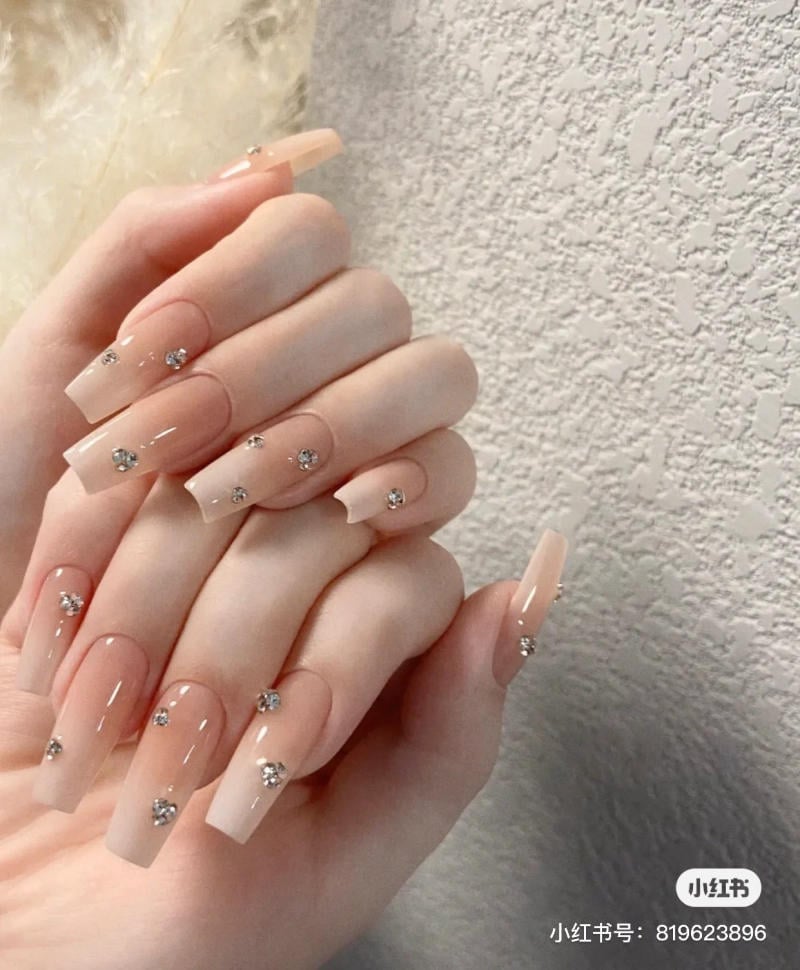 Bàn tay với móng tay dài, màu nude, trang trí đá lấp lánh, nền tường trắng.