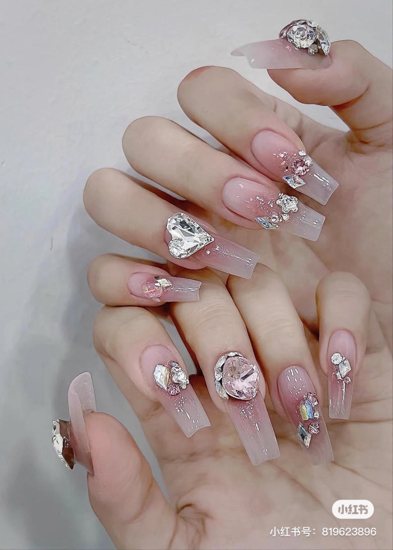 Bàn tay với móng tay dài, trang trí đá quý và kim tuyến lấp lánh trên nền hồng nhạt.