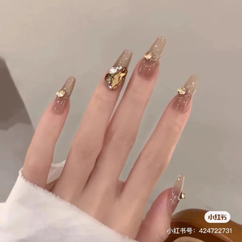 Bàn tay với móng tay dài, được trang trí bằng lớp sơn màu nâu nhạt và đính đá, tạo vẻ sang trọng.