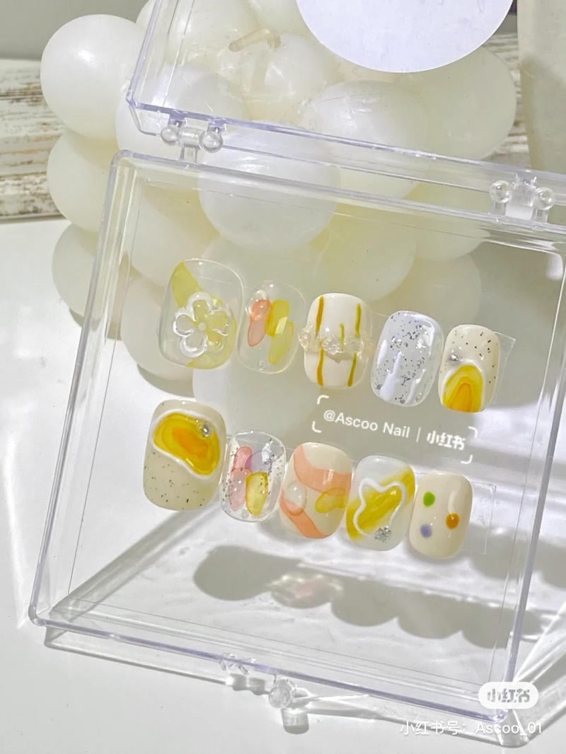 Bộ móng giả với họa tiết đa dạng, màu sắc tươi sáng, đặt trong hộp nhựa trong suốt trên nền trắng.