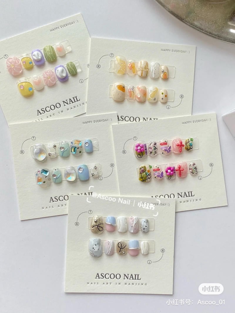 Bộ móng tay giả nhiều màu sắc và họa tiết khác nhau được trưng bày trên các tấm thẻ trắng.