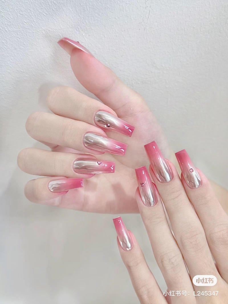 Bàn tay với móng tay dài được sơn ombre hồng và bạc, trang trí bằng đá nhỏ lấp lánh.