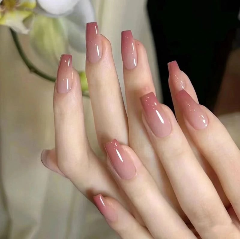 Bàn tay với móng dài được sơn màu hồng ombre bóng, nền phía sau có hoa trắng nhạt.