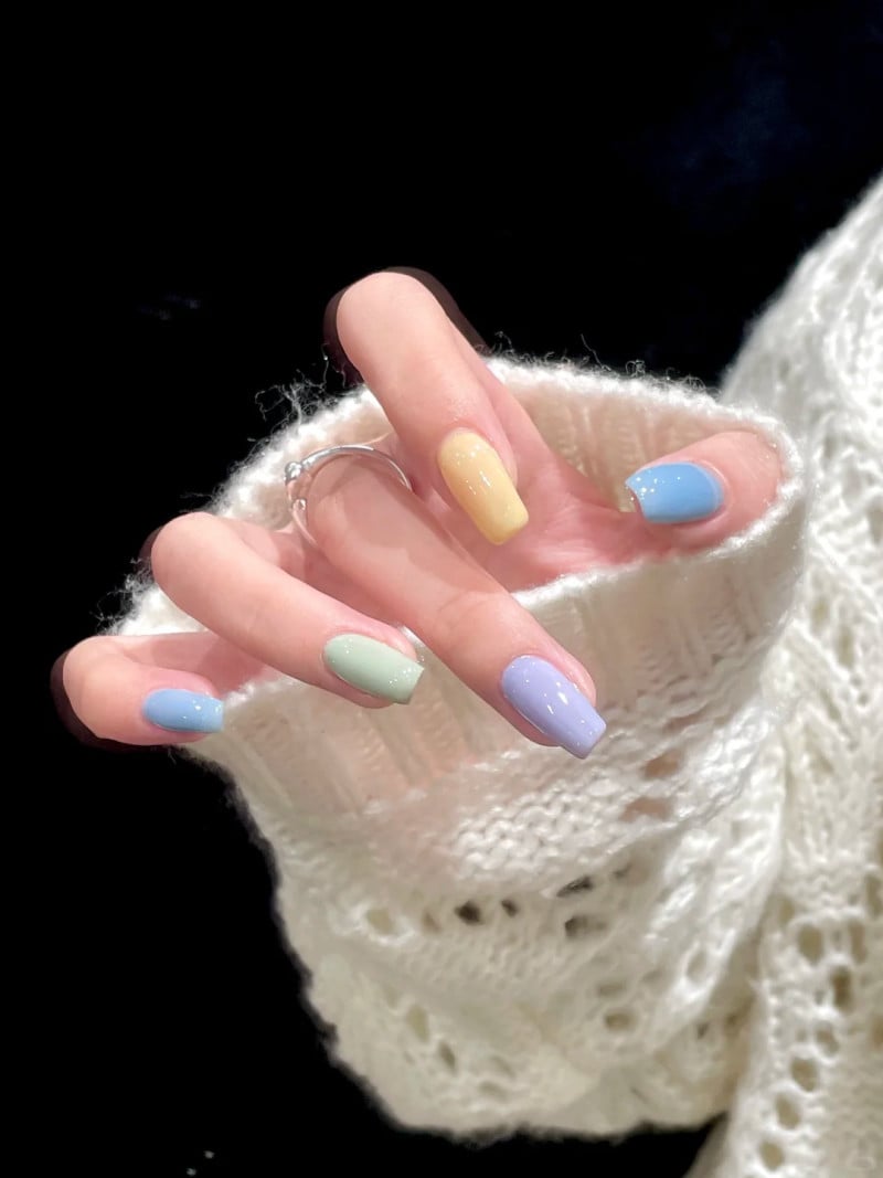 Bàn tay với móng tay sơn nhiều màu pastel, đeo nhẫn bạc, áo len trắng, trên nền đen.