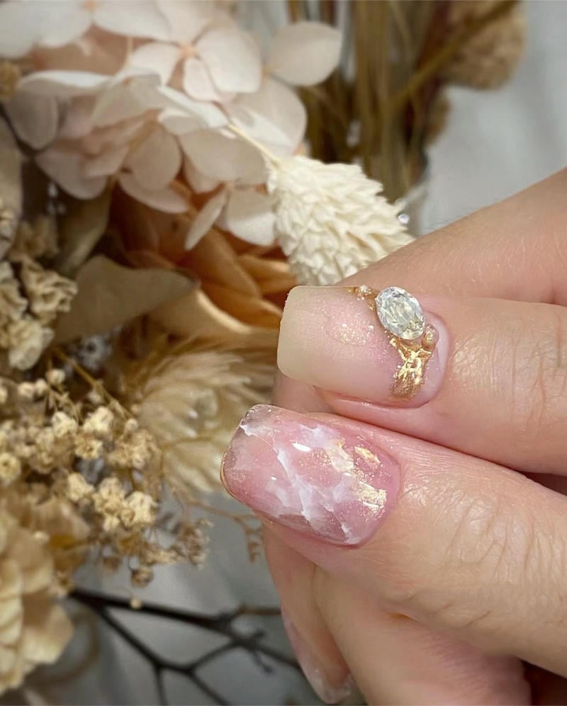 nail-van-da-dep__10__8def379a021d49cda8d384e9069e53a4 Bàn tay với móng tay được trang trí nghệ thuật, nổi bật với nhẫn có đá quý, nền là hoa khô trang trí.