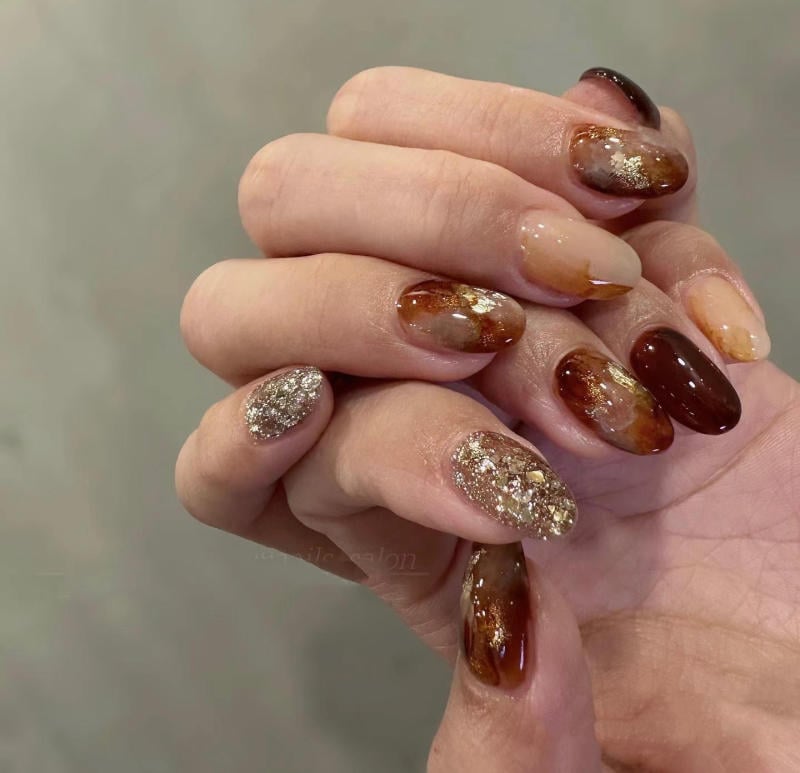 nail-van-da-dep__14__4a208dd3bd264ed4ac7bdd95e5df9e5a Bàn tay với móng tay được trang trí bằng sơn màu nâu và vàng lấp lánh, tạo kiểu nghệ thuật.