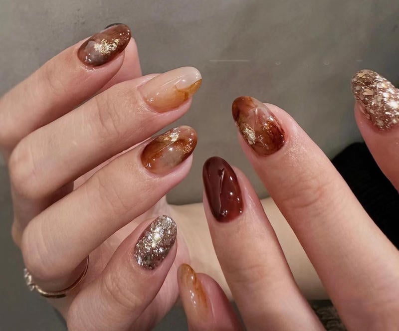 nail-van-da-dep__15__8d25492f9236418bbcea61fa246b8d17 Bàn tay với móng tay được sơn màu nâu và vàng ánh kim, thiết kế đa dạng và nghệ thuật.