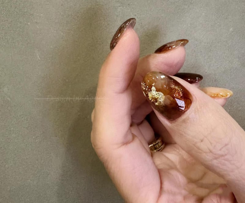 nail-van-da-dep__19__7bd7f619669d40f3a486ef4a8ef91149 Bàn tay với móng tay sơn màu nâu bóng, có họa tiết vàng lấp lánh trên nền xám.