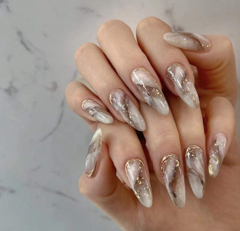 nail-van-da-dep__20__037bf980e3b2405c95b0718e17d142f9 Bàn tay với móng tay dài được trang trí họa tiết cẩm thạch và viền vàng trên nền trắng.