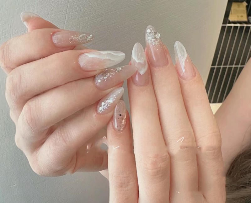 nail-van-da-dep__22__a66031e09f214e2fb7594f7ddf8f318a Bàn tay với móng tay dài được sơn màu trắng và ánh kim, có họa tiết trang trí lấp lánh.