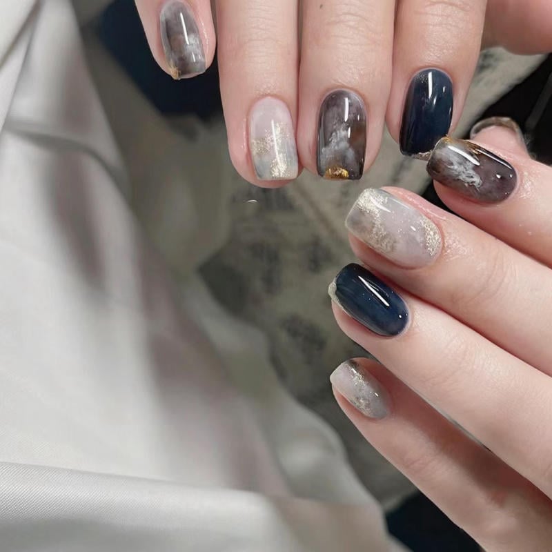 nail-van-da-dep__25__18b5b5242dd649dabcd8810a2ff35e2b Bàn tay với móng tay được sơn nghệ thuật, phối màu xanh đen và xám, có họa tiết vân đá và ánh kim.