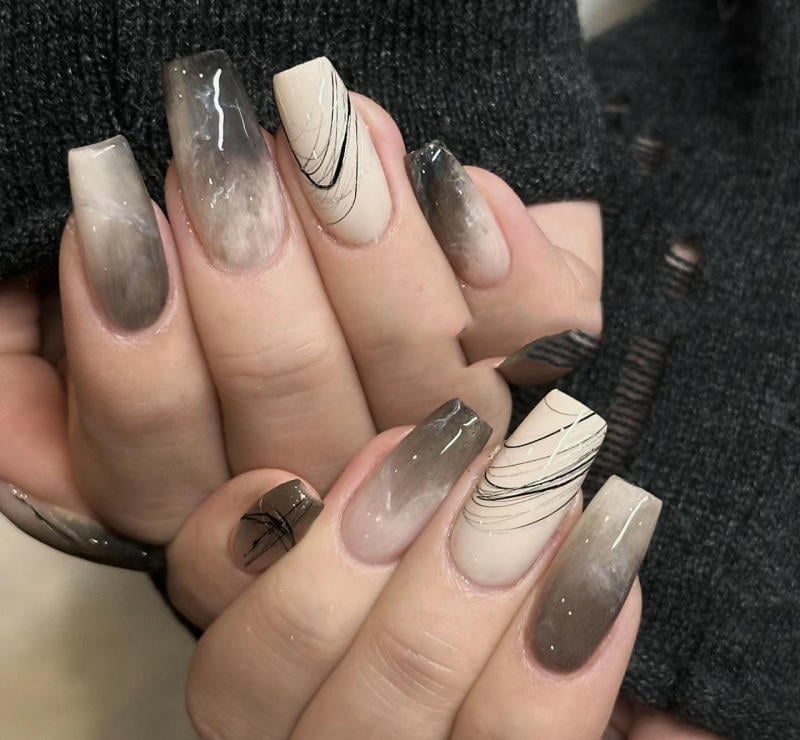 nail-van-da-dep__27__a290b4e438854814a8d33bd75112f1e1 Bàn tay với móng tay dài, được sơn màu ombre và trang trí họa tiết đường kẻ đen trên nền màu be.
