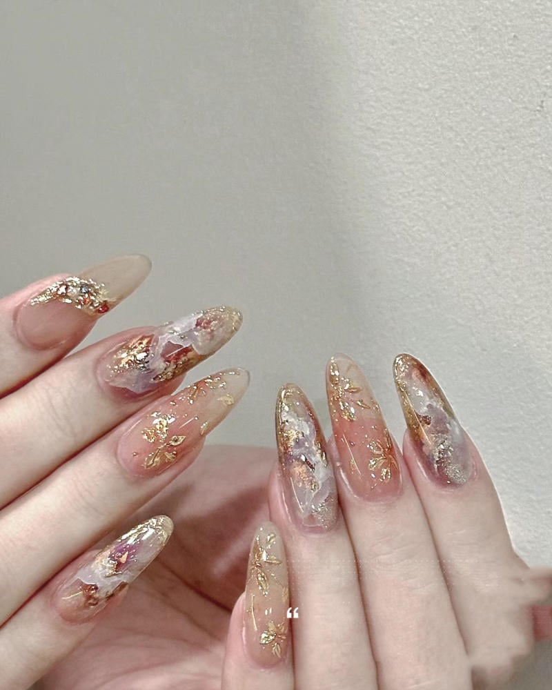 nail-van-da-dep__2__463db00c7c5f4bf5a40292d076b925ea Bàn tay với móng tay dài được trang trí bằng họa tiết vàng và trắng trên nền màu hồng nhạt.