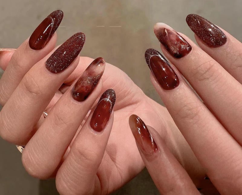 nail-van-da-dep__30__a1785d117fbe421191484086c2966003 Bàn tay với móng tay được sơn màu đỏ nâu, có họa tiết vân đá và lấp lánh.