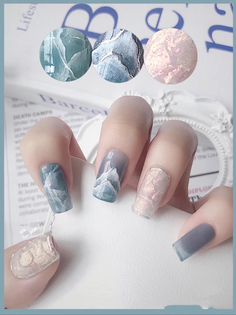nail-van-da-dep__34__80785d4e8b724e85a5c854517bc25d6f Bàn tay với móng tay được sơn họa tiết đá cẩm thạch, màu sắc xanh và hồng nhạt, nền là trang sách.