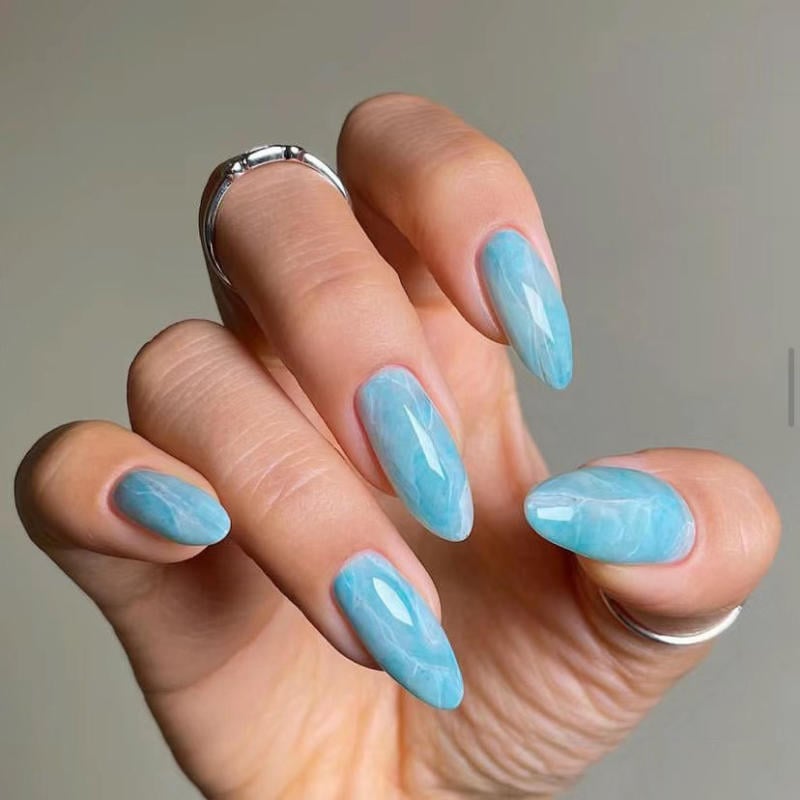 nail-van-da-dep__35__fe14961a2c804e2e8a21d648a8925d56 Bàn tay với móng tay giả màu xanh, được đánh bóng kỹ lưỡng, đeo nhẫn bạc trên ngón tay giữa.