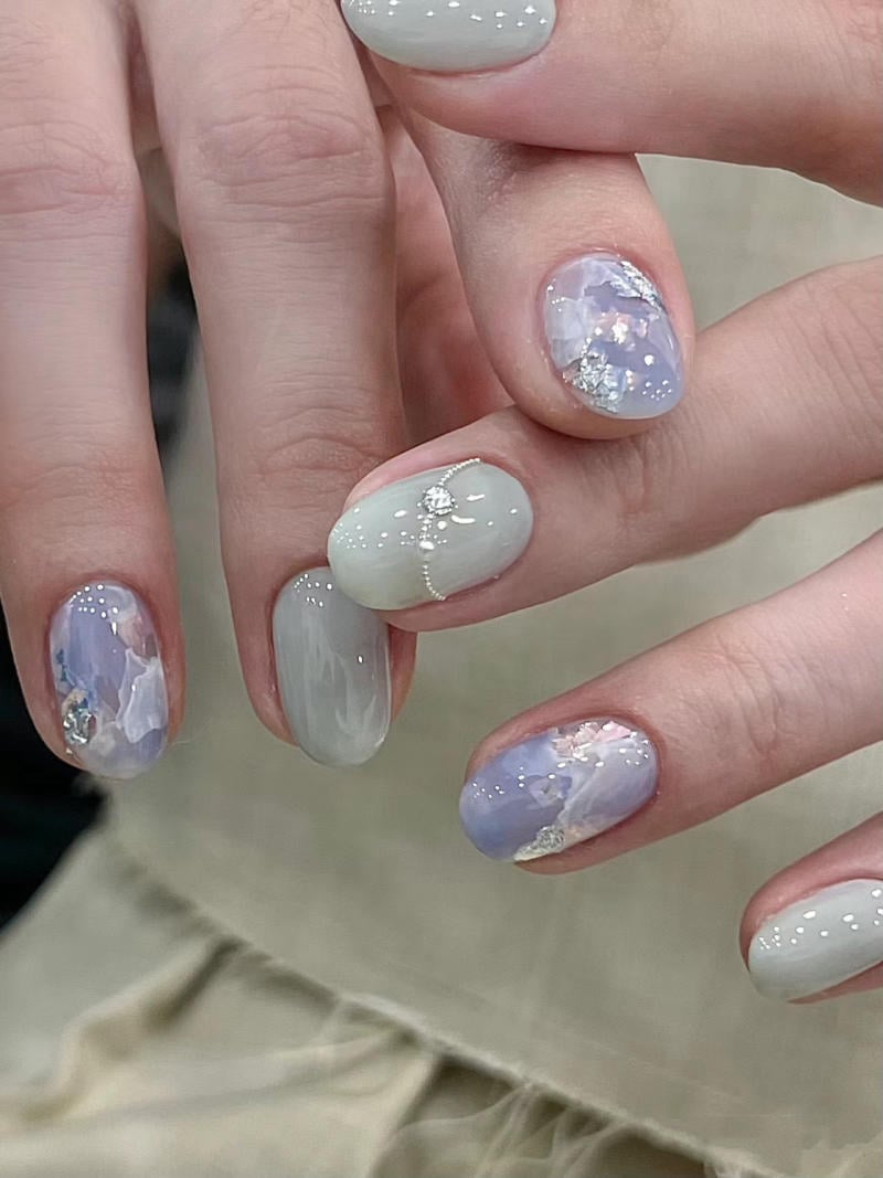 nail-van-da-dep__36__e255199b0d0441879b094f2142c03a6d Bàn tay với móng tay được sơn màu xanh nhạt và tím, trang trí hoa văn lấp lánh và họa tiết trừu tượng.