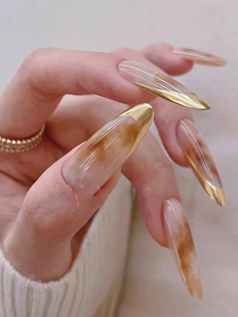 nail-van-da-dep__38__50ddc57a72c2485e94feb49699807e89 Bàn tay với móng dài, thiết kế họa tiết nâu và vàng, mang trang sức vòng tay.