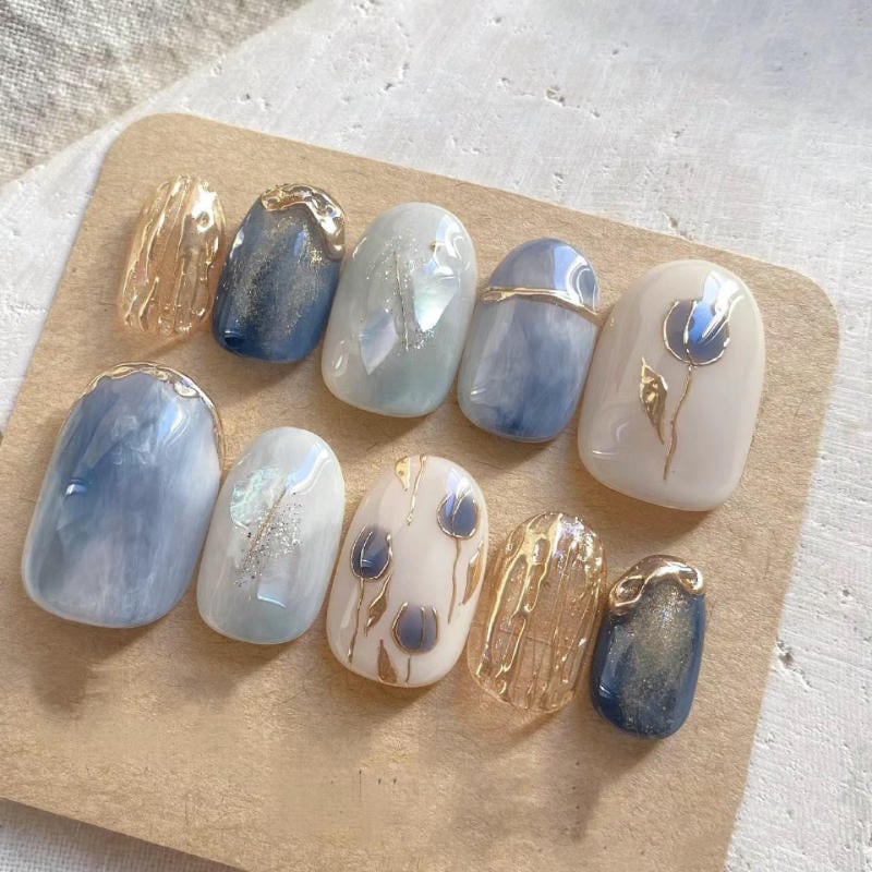 nail-van-da-dep__39__fed7edd5f9dc49f4b23ee579ca3cc628 Bộ móng giả với thiết kế màu xanh và trắng, trang trí họa tiết vàng và hoa văn tinh tế trên nền vải trắng.