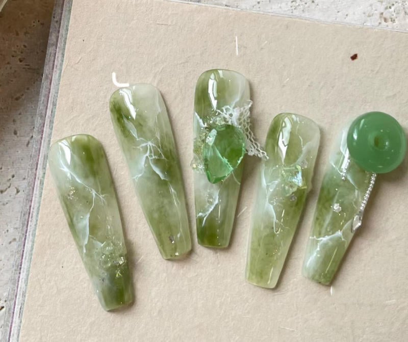 nail-van-da-dep__45__2bc6e48d3d6d40d7a05acd6de81481e1 Bộ móng tay giả màu xanh ngọc, có hoa văn và đính đá trang trí cầu kỳ, đặt trên bề mặt màu be.