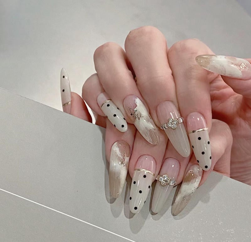 nail-van-da-dep__53__1fdf6e88042945b6ad87b5437dbc5b07 Bàn tay với móng dài được trang trí họa tiết chấm bi và nhũ, kết hợp nhẫn hoa nhỏ trên ngón tay.