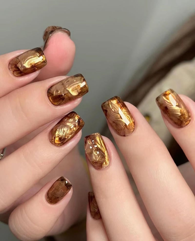 nail-van-da-dep__57__a0a87f49cd2a4a14aa06097028210850 Bàn tay với móng tay được sơn kiểu họa tiết vân đá màu nâu vàng, bóng và sáng.