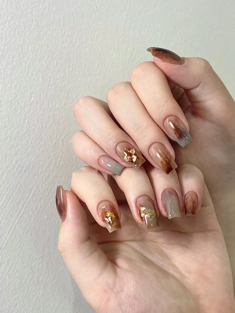 nail-van-da-dep__5__d592921b18054c828f5f625f5b326bb2 Bàn tay với móng tay được sơn màu nâu và xám, trang trí họa tiết lá vàng lấp lánh.