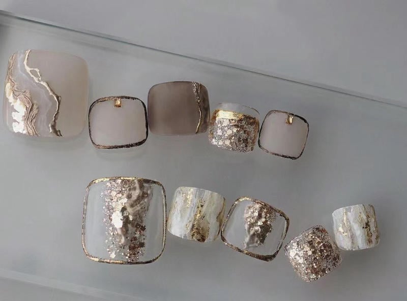 nail-van-da-dep__62__63c0a5ab63b94292bdd1c28033540384 Bộ móng giả hình vuông với họa tiết màu nâu và ánh kim, được sắp xếp trên bề mặt trong suốt.