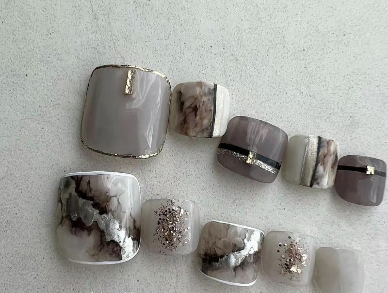 nail-van-da-dep__64__4f5f7295d81040088a161013c6f95aae Bộ móng tay giả màu xám và bạc, thiết kế đa dạng với các chi tiết vân đá và lấp lánh trên nền trắng.