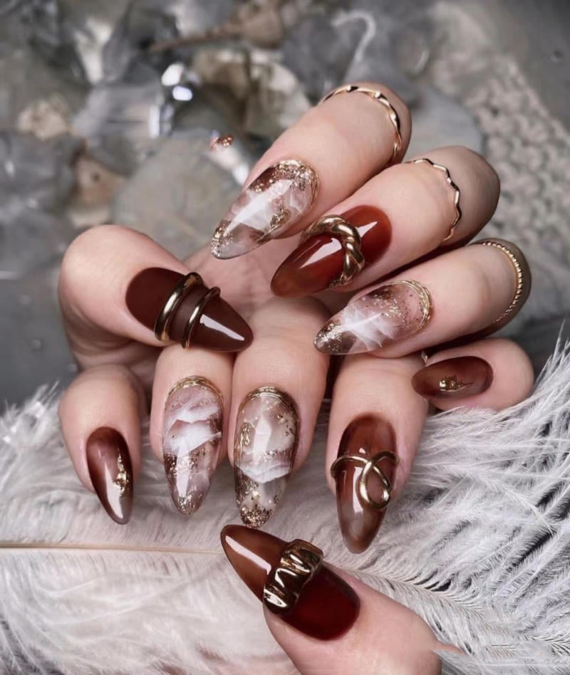 nail-van-da-dep__73__35fe72c28a4545aa899eb923cf92ad20 Bàn tay với móng tay dài, sơn màu nâu và hoa văn trang trí tinh tế, bên dưới là lông vũ màu trắng.