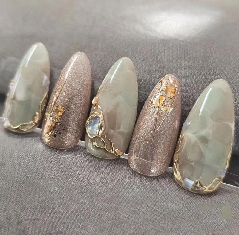 nail-van-da-dep__74__1daae67a7cdd442ab5955a8f3aa79d8b Bộ móng tay giả màu xám và nâu, trang trí hoa văn vàng và đá lấp lánh trên nền vải xám.