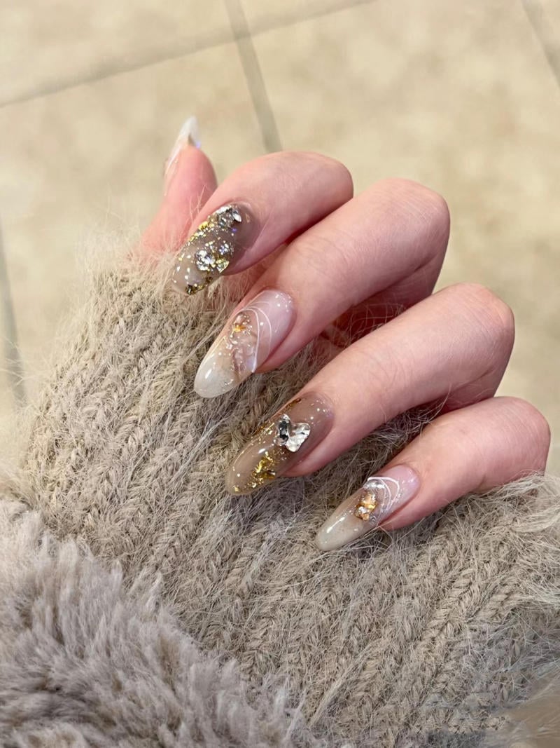 nail-van-da-dep__75__b0ab6166e7cb4e4c812239debf78cfce Bàn tay với móng tay dài được trang trí hoa văn và kim tuyến, mặc áo len màu xám.