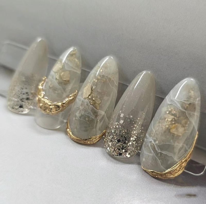 nail-van-da-dep__78__16122838787f4c05a59820a507531ec7 Bộ móng tay giả màu xám trắng với họa tiết ánh kim và trang trí vàng, bày trên nền vải màu xám nhạt.