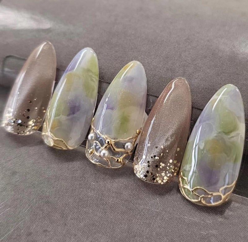 nail-van-da-dep__79__deadd1dfcb4b487791e9c7dcd9660885 Móng tay giả dài với thiết kế màu sắc và họa tiết ánh kim, đính đá và ngọc trai trên nền vải xám.