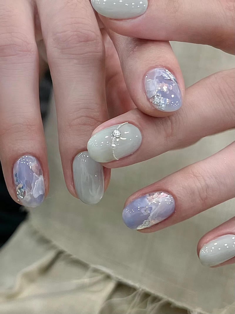 nail-van-da-dep__80__c7a5a79dd57c422a961b84cdc5372008 Bàn tay với móng tay được sơn màu tím và xám, trang trí hoa văn tinh tế và lấp lánh.