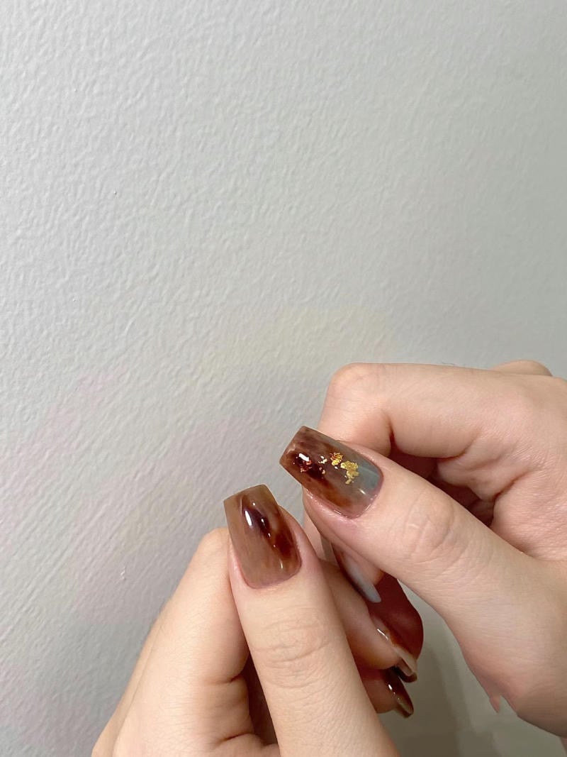 nail-van-da-dep__9__4240a2c3029f44d985be64a8ed4ae27f Hai bàn tay với móng tay dài, sơn màu nâu và họa tiết vàng nhỏ trên nền tường màu trắng.